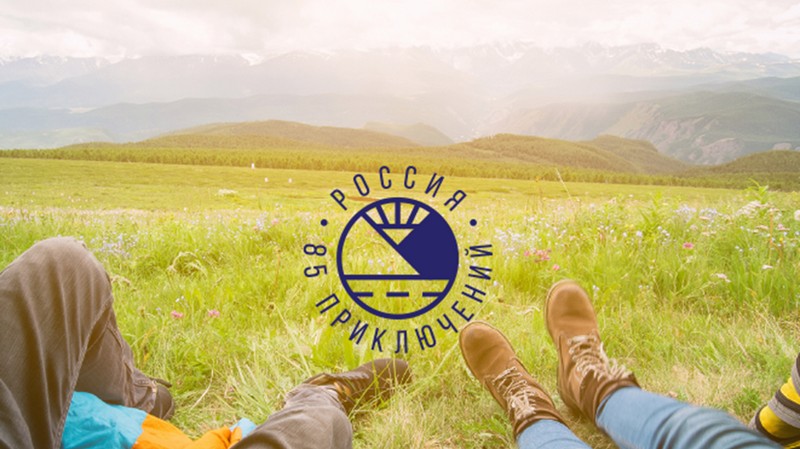 на изображении фон зеленой травы и по центру на нем нанесен логотип проекта с синей надписью: Россия, 85 приключений